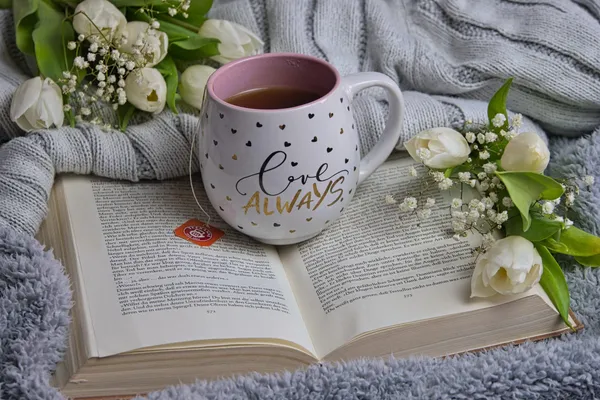 Teetasse auf aufgeschlagenem Buch mit weißen Tulpen auf grauer Strickdecke