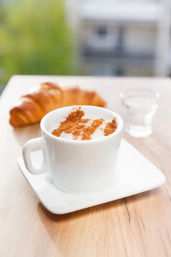 Cappuccino mit Zimt-Schaum neben Croissant und Wasserglas